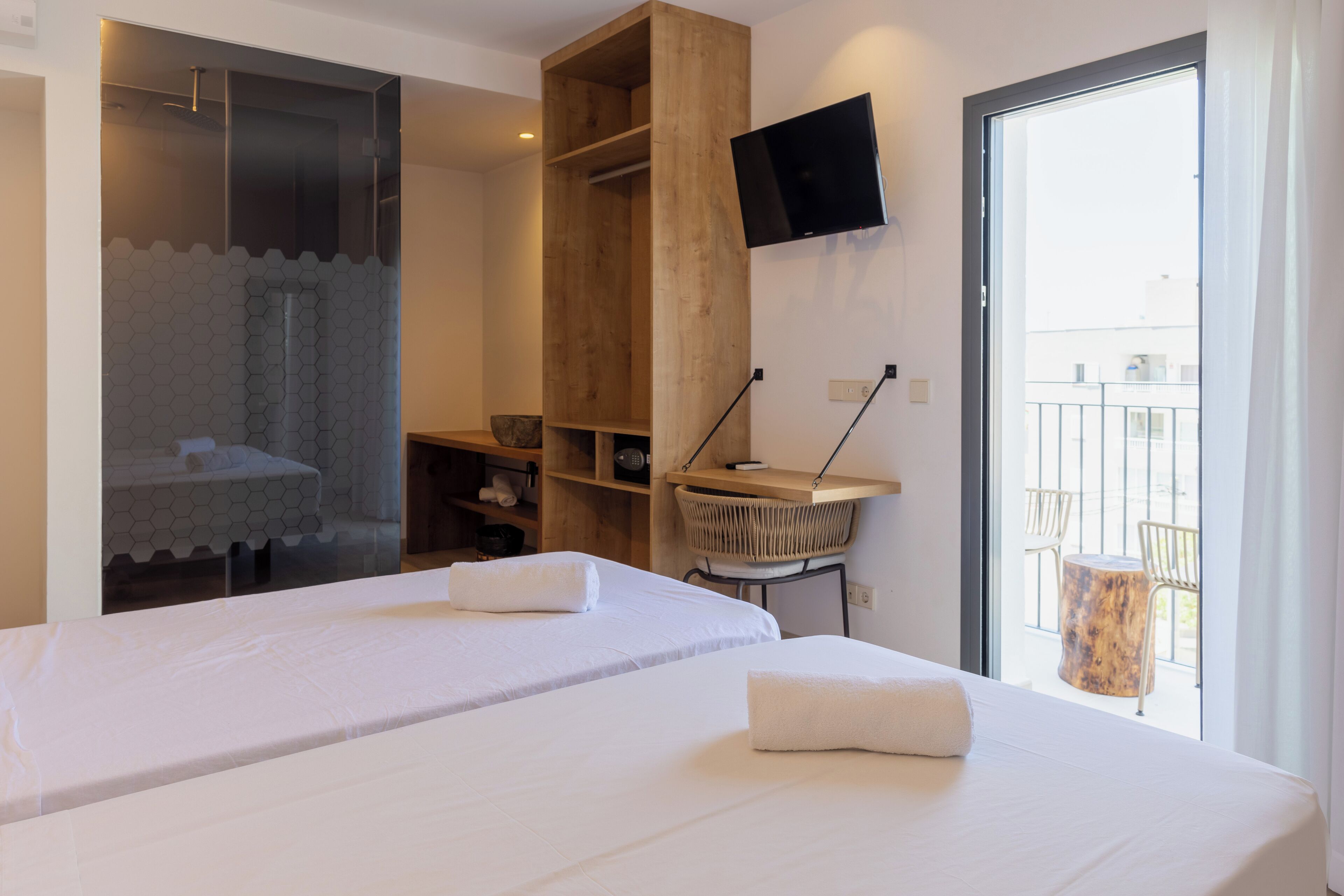 A hotel room with two beds, a shower area, a TV mounted on the wall, and a balcony with chairs.