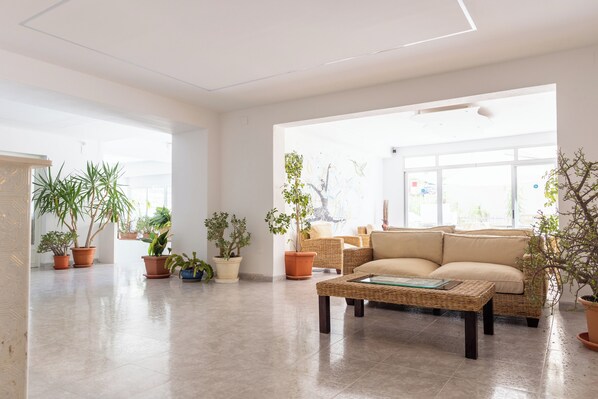 A spacious living room with a beige sofa, a wicker coffee table, and several potted plants.