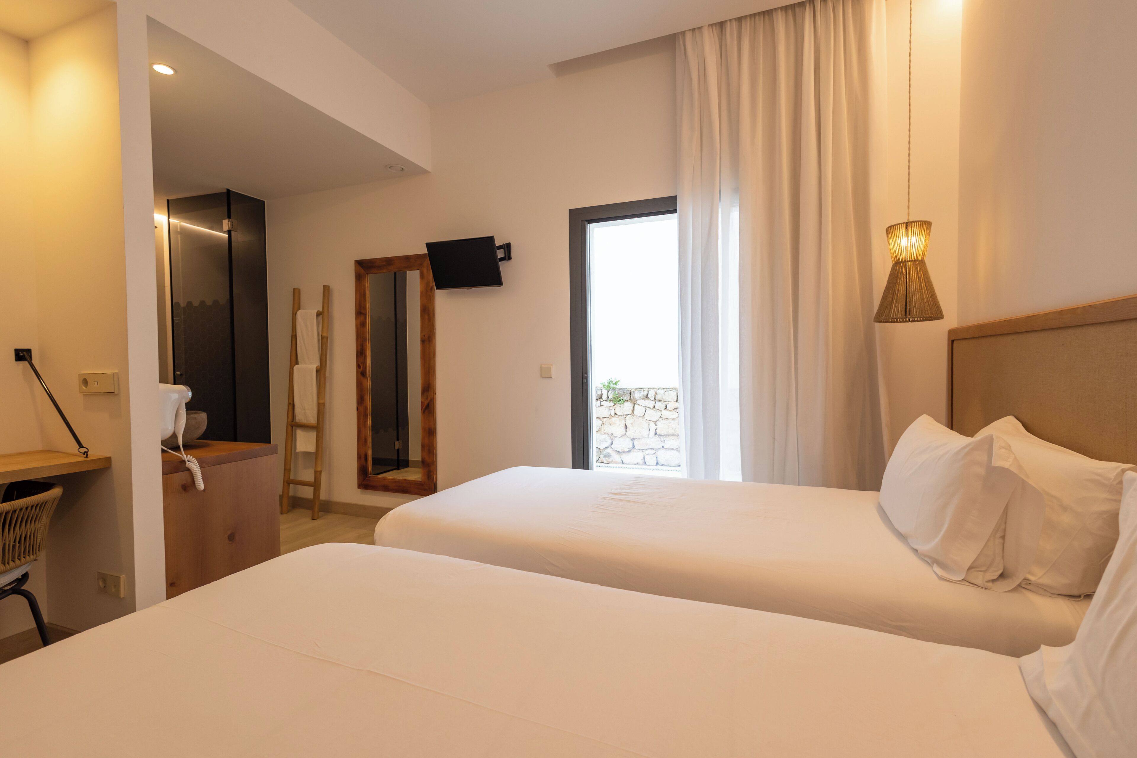 Quarto de hotel com duas camas, uma escrivaninha de madeira, um espelho, uma televisão e uma janela com cortinas.