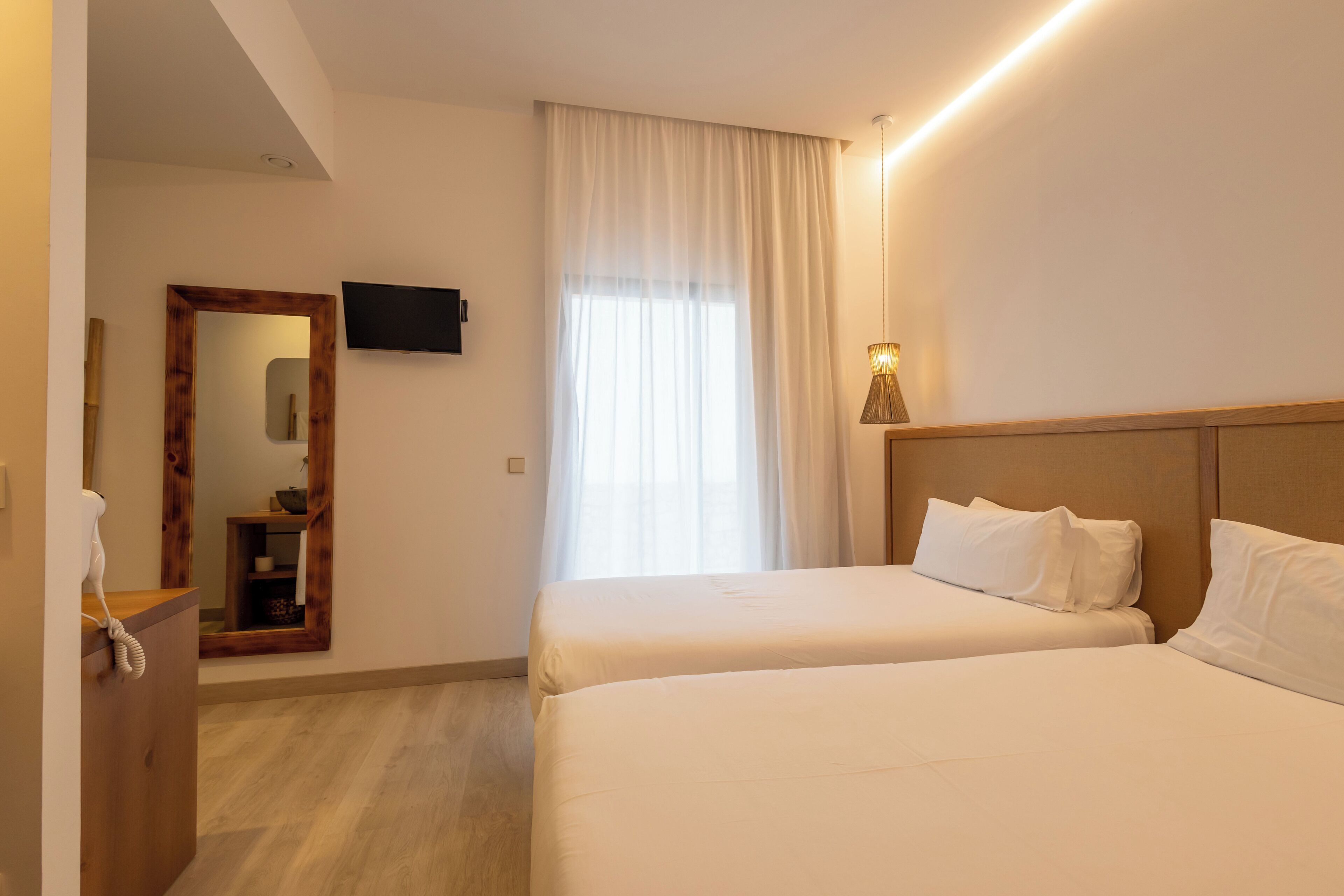 A hotel room with two beds, a TV mounted on the wall, a desk with a telephone, and a window with curtains.