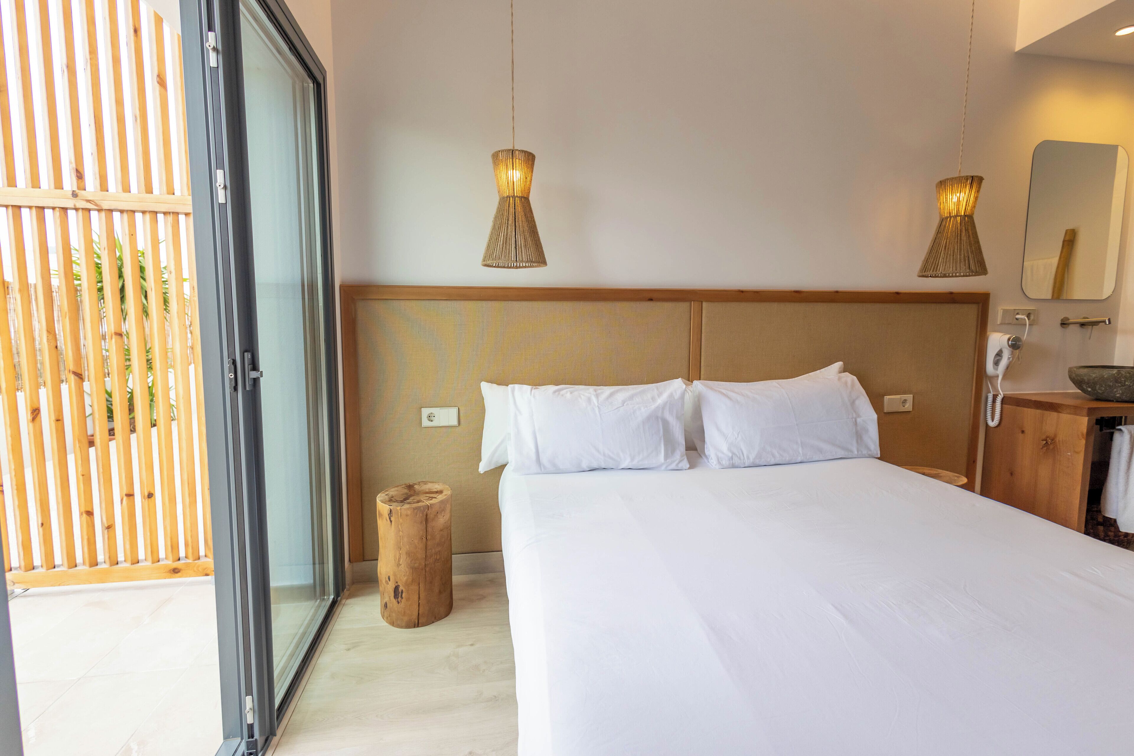 A bedroom with a large bed, wooden headboard, two pendant lights, a wooden stool, and a sliding glass door leading to an outdoor area.