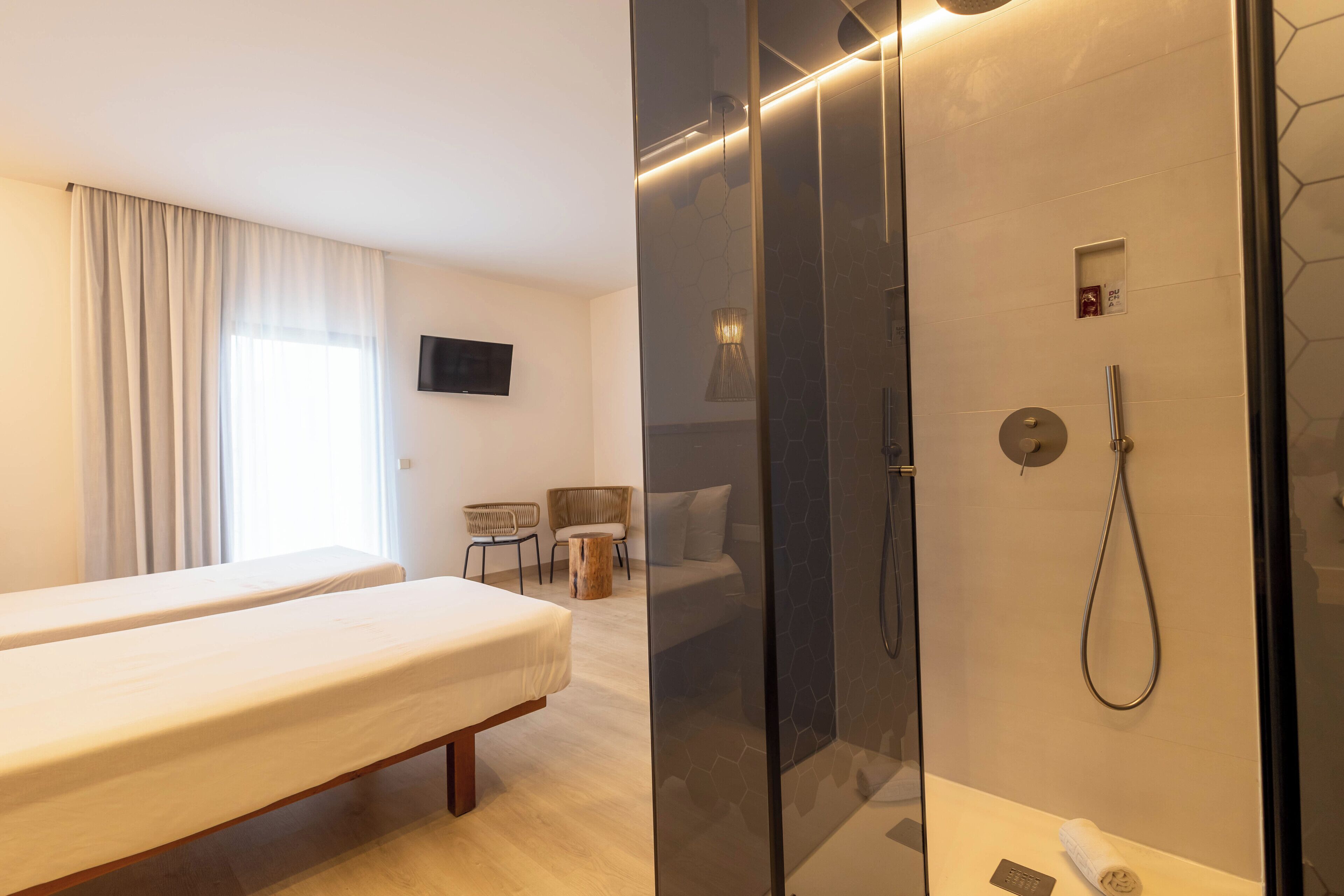 Habitación de hotel con cama, zona de ducha, televisor y espacio de descanso con sillas.