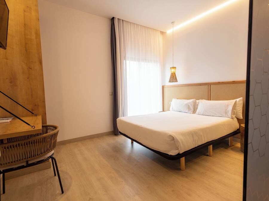 Una habitación de hotel moderna con una cama, un escritorio de madera, una silla de mimbre y una lámpara de noche.
