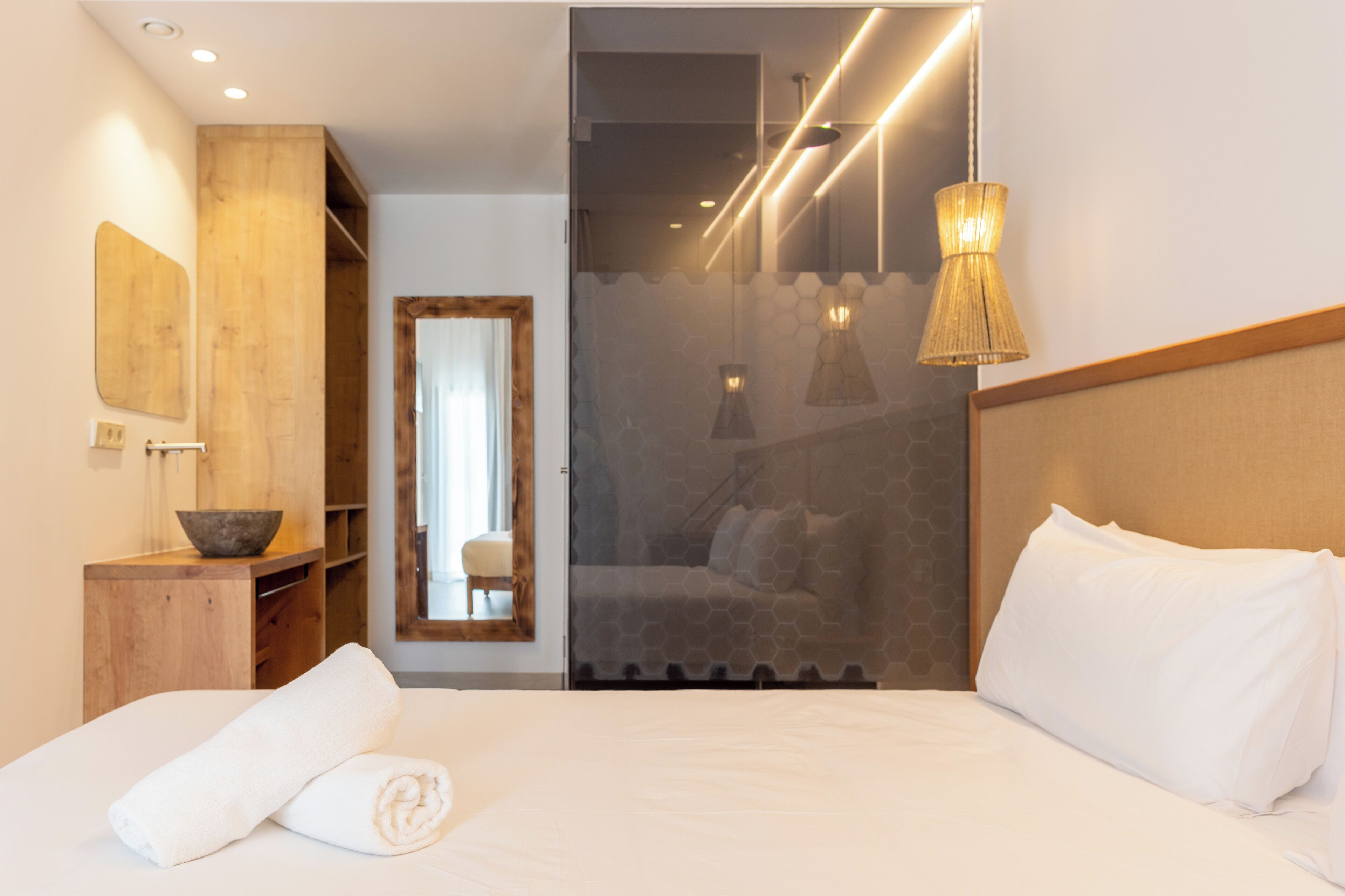 Una habitación de hotel moderna con una cama grande, una mesita de madera con un cuenco, un espejo y un panel de vidrio que conduce a un baño.
