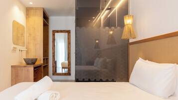 Una habitación de hotel moderna con una cama grande, una mesita de madera con un cuenco, un espejo y un panel de vidrio que conduce a un baño.