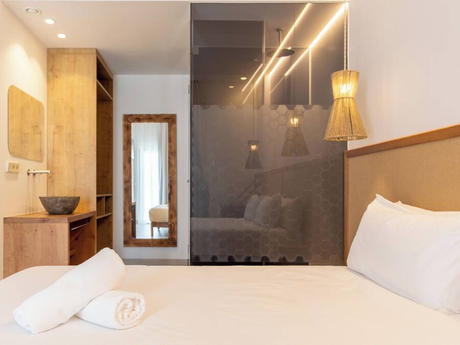 Una habitación de hotel moderna con una cama grande, una mesita de madera con un cuenco, un espejo y un panel de vidrio que conduce a un baño.