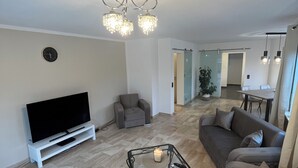 A modern living room with a flat-screen TV, a glass coffee table with a candle, a sofa, a chair, and a ceiling light fixture.