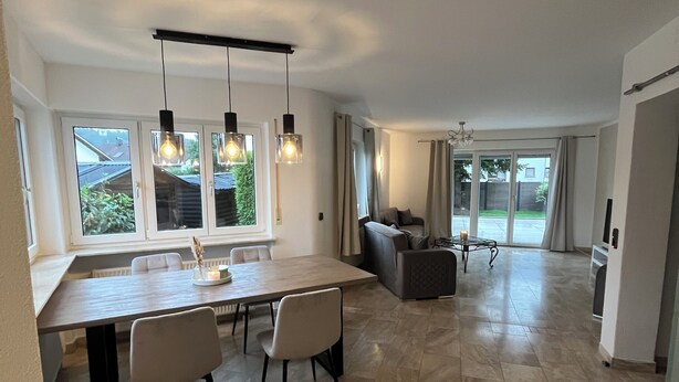 A spacious living area with a dining table, chairs, a sofa, and a large window providing a view of the outdoors.