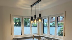 A dining area with a large table, four chairs, pendant lights, and a view through large windows.
