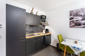 Une cuisine de taille réduite avec des meubles de couleur sombre, un four à micro-ondes, une machine à café et un évier. On y trouve une table à manger recouverte d’une nappe blanche et une chaise verte.