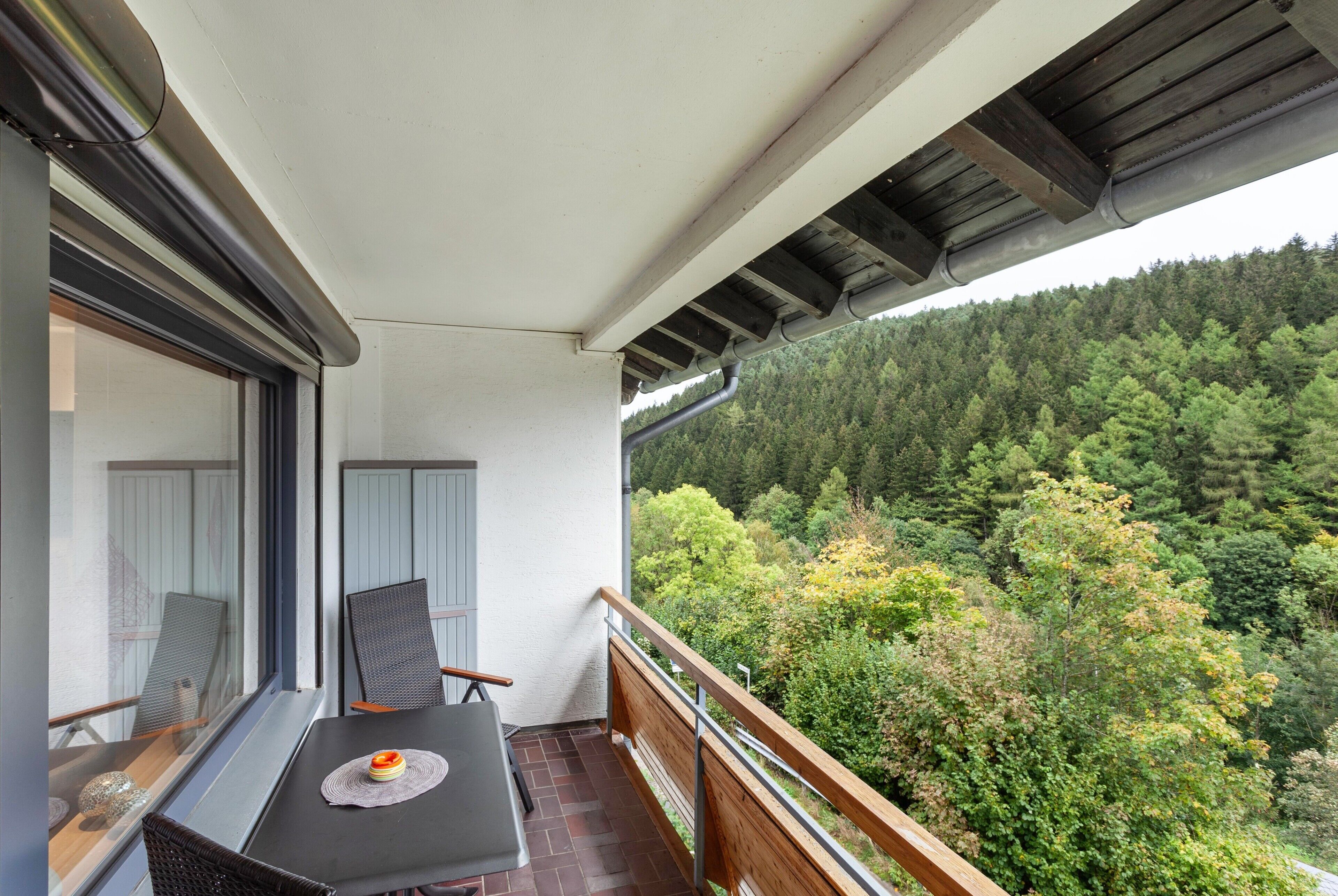 A balcony with a table and chairs, overlooking a forested hillside.