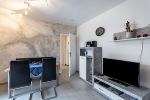 Un salon moderne doté d’un téléviseur à écran plat, d’un coin repas avec deux chaises, d’un meuble blanc et d’une étagère ornée d’objets décoratifs.