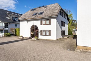 Ein zweistöckiges Gebäude mit geneigtem Dach, einem zentralen Eingang mit einer Tür und einem kleinen Gartenbereich mit Topfpflanzen.
