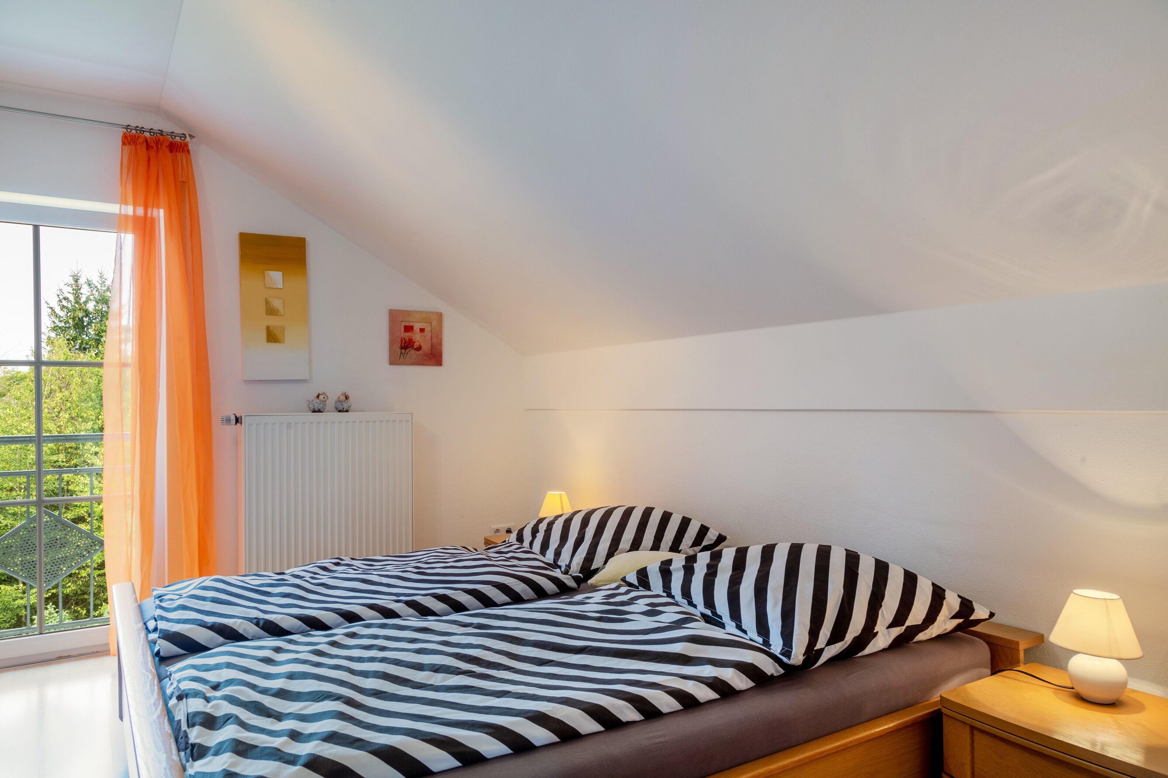 A bedroom with a bed, nightstands, a lamp, a window with curtains, and a radiator.