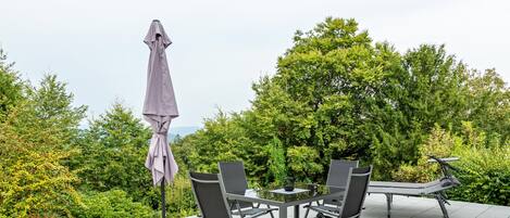 An outdoor patio with a table and chairs, surrounded by greenery and a view of trees.
