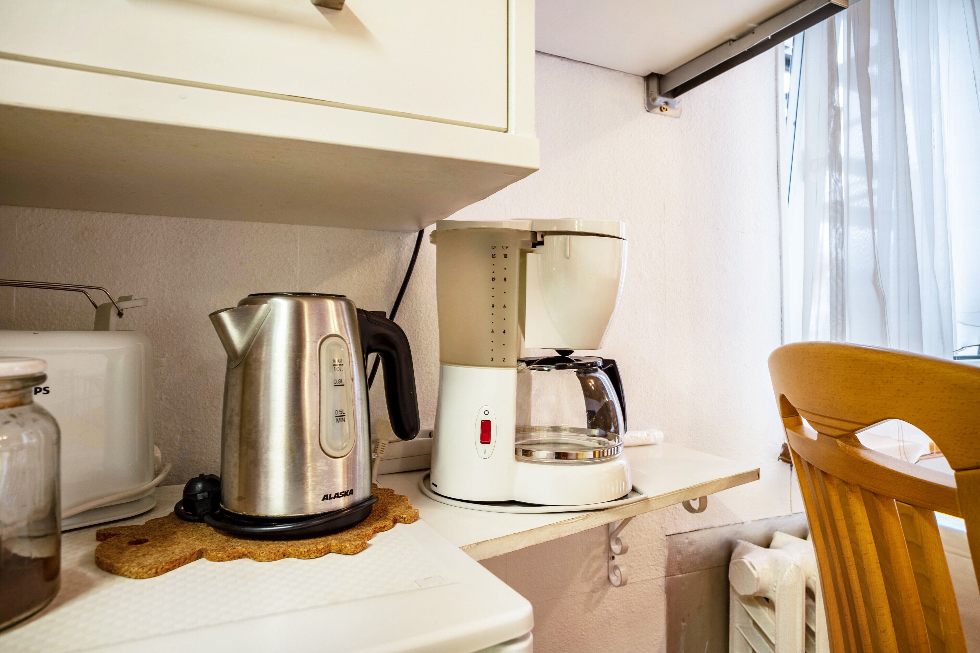 A kitchenette with a kettle, coffee maker, toaster, and a wooden chair.