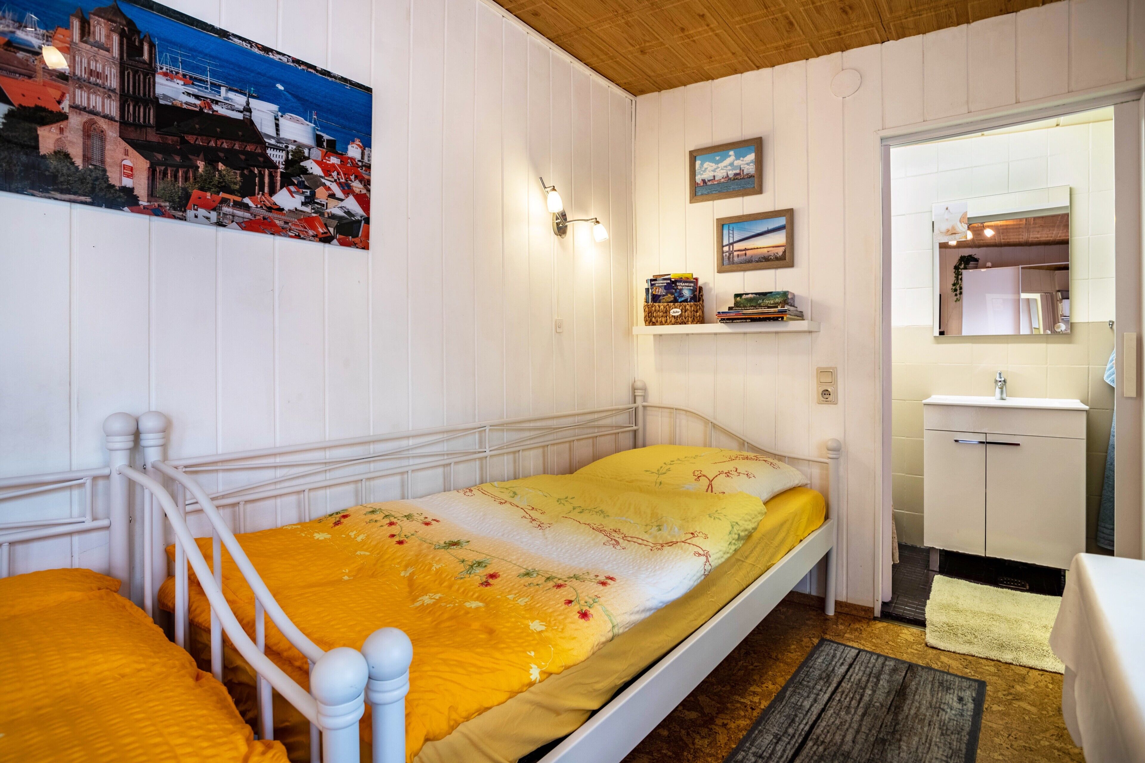 A single-bed room with a wooden ceiling, a white wall with a framed picture, a small shelf with books, and a white bathroom with a mirror and sink.