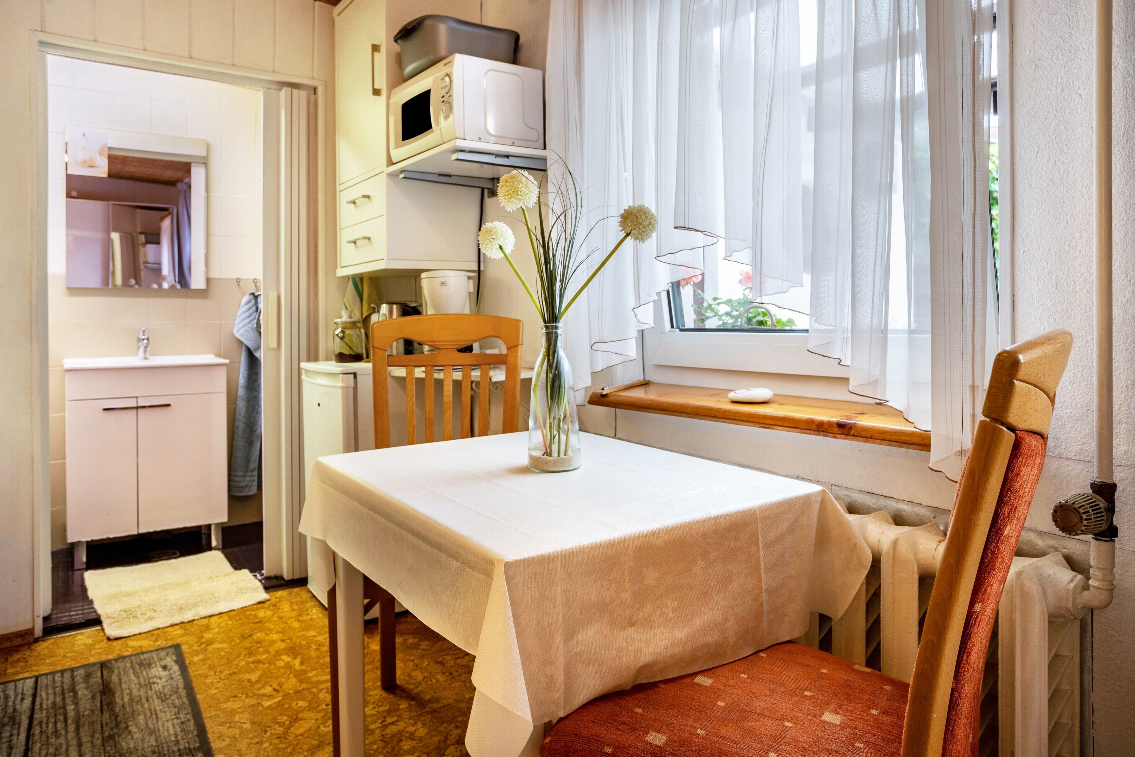 A small kitchen with a table, chairs, a vase with flowers, a window with curtains, and a microwave above the cabinets.