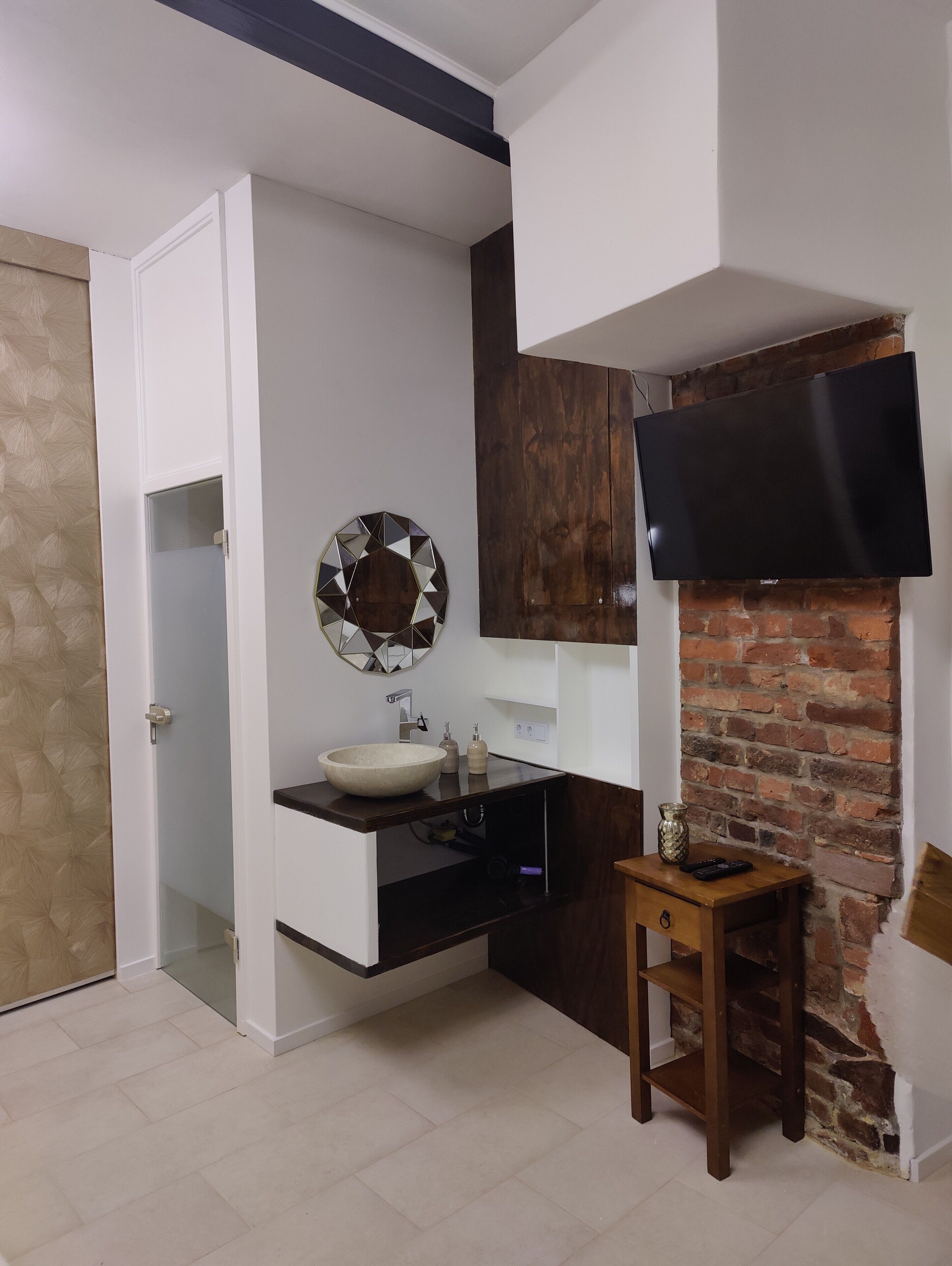 Un baño moderno con un lavamanos blanco, un espejo redondo, un televisor de pantalla plana montado en una pared de ladrillo y una mesa de madera con un florero y controles remotos.