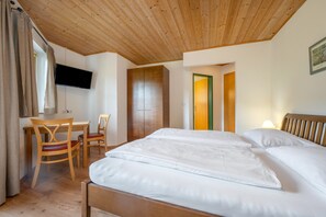 A hotel room with a wooden ceiling, a bed with white bedding, a desk with a chair, and a television mounted on the wall.