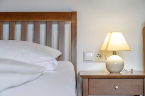 A neatly made bed with white linens, a wooden headboard, a bedside table with a lamp, and a power outlet.