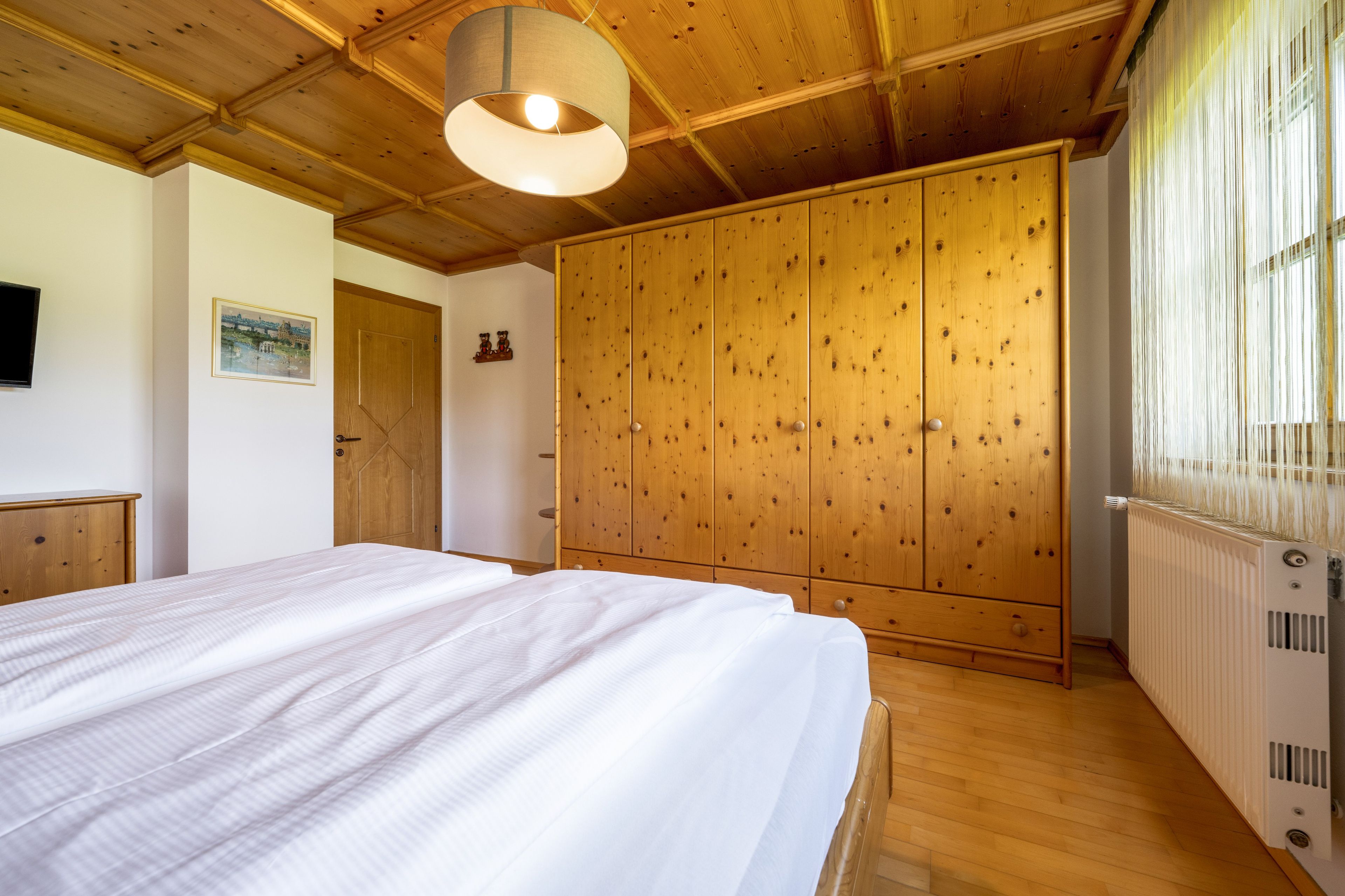 A bedroom with a wooden ceiling, a bed with white bedding, a wardrobe, and a radiator.