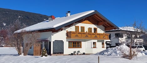 A two-story chalet with a sloped roof, wooden balcony, and a snow-covered landscape.