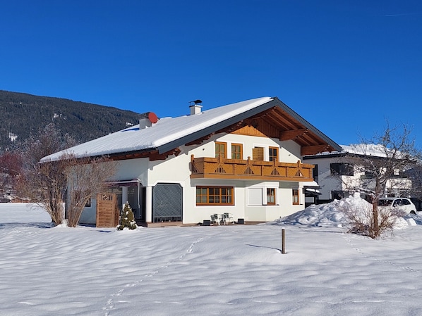 A two-story chalet with a sloped roof, wooden balcony, and a snow-covered landscape.