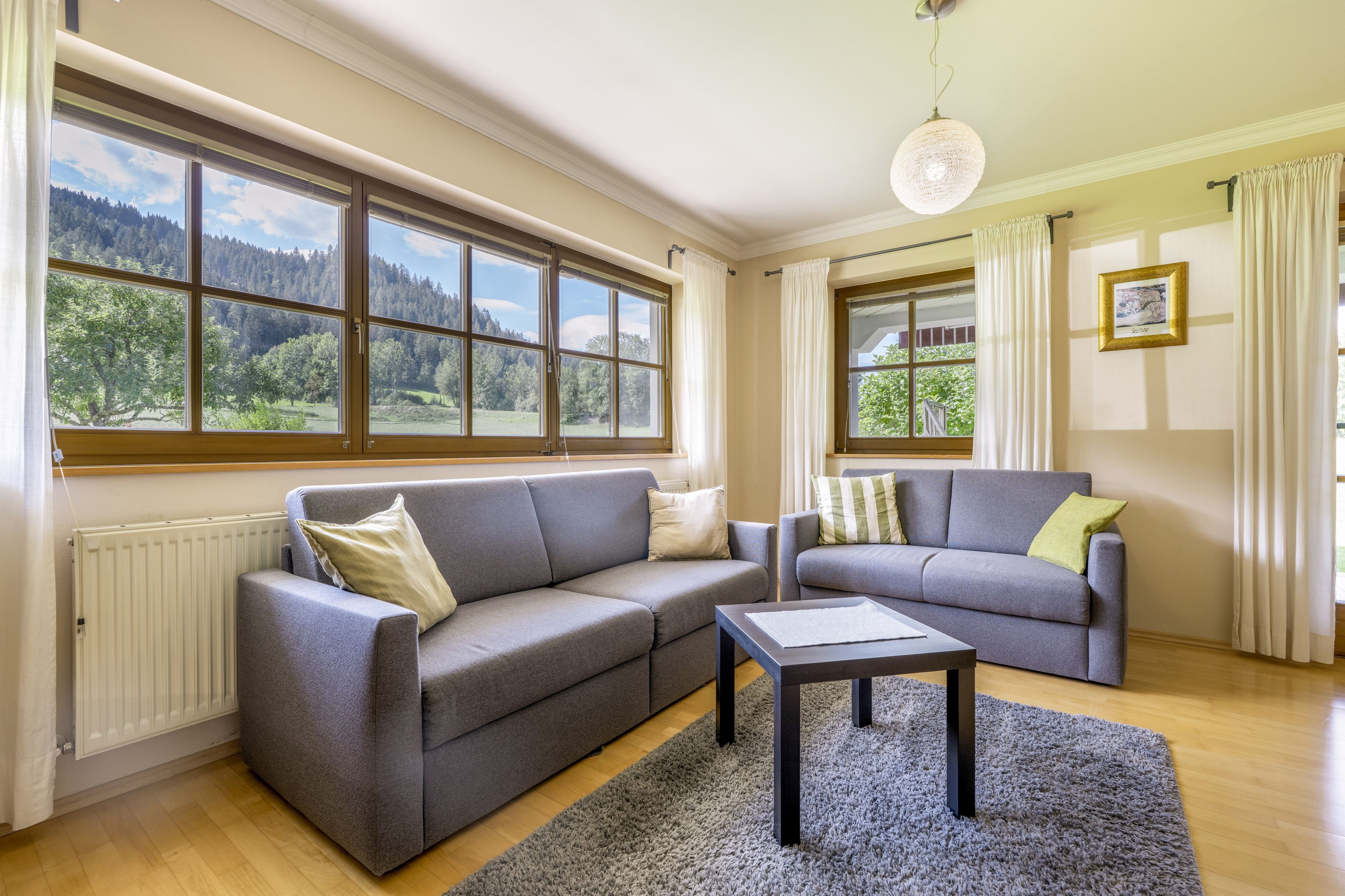 A modern living room with a grey sofa set, a coffee table, and large windows offering a view of a forested landscape.