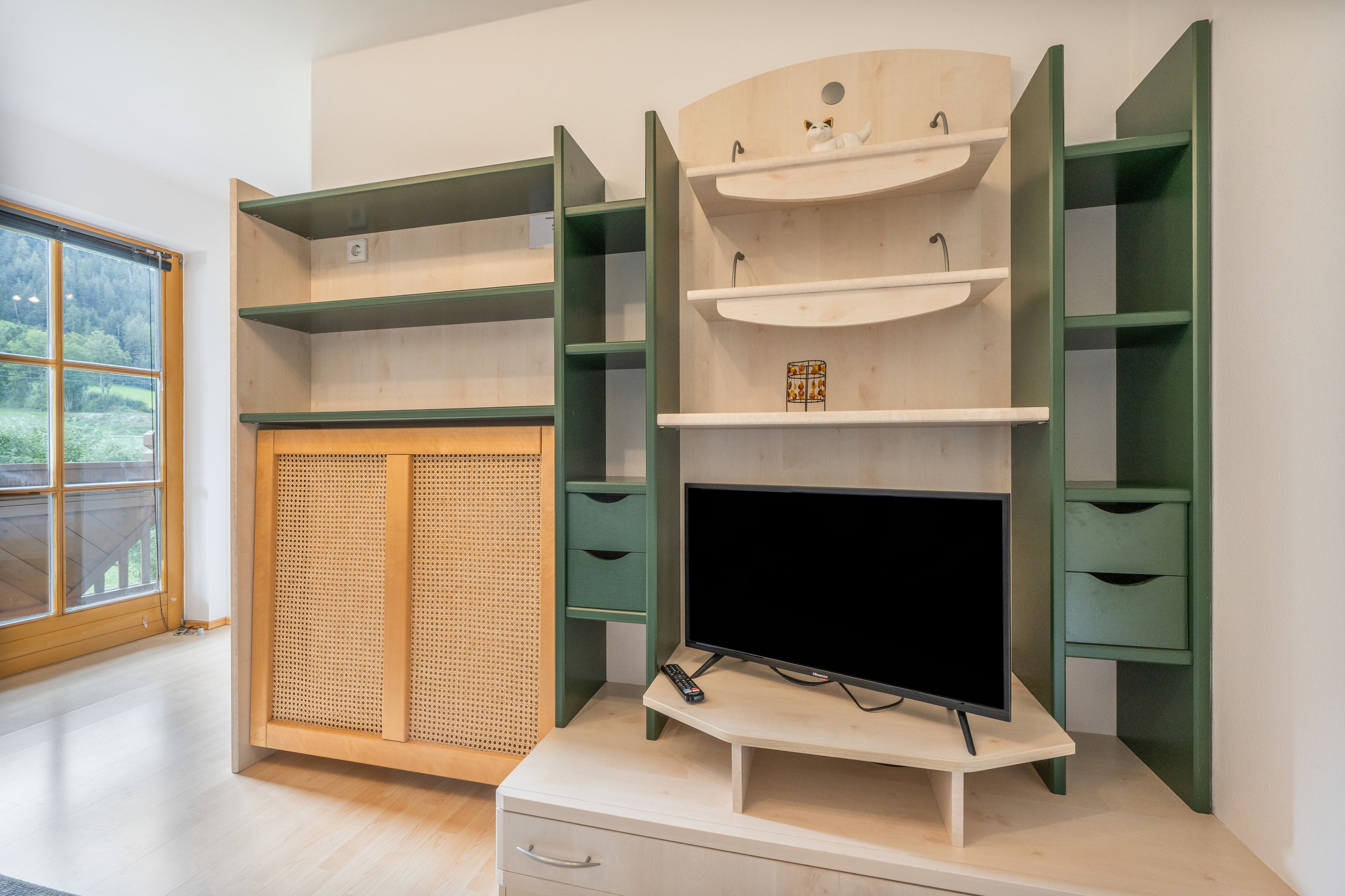 A room with a built-in TV on a stand, a wooden cabinet with a mesh screen, and a window with a view of trees.