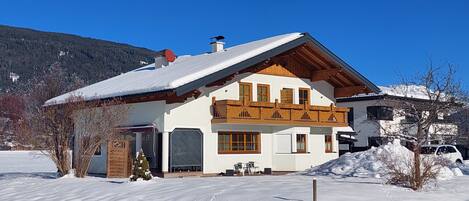 A two-story chalet with a sloped roof, wooden balcony, and a snow-covered landscape.