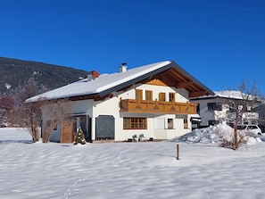 A two-story chalet with a sloped roof, wooden balcony, and a snow-covered landscape.