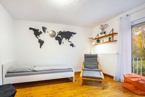 A room with a bed, a chair, a world map on the wall, a shelf with decorative items, and a window with a view of trees.