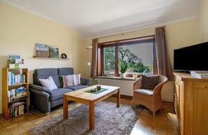 Ein Wohnzimmer mit einer Couch, einem Couchtisch, einem Bücherregal, einem Fernseher und einem Fenster mit Vorhängen.