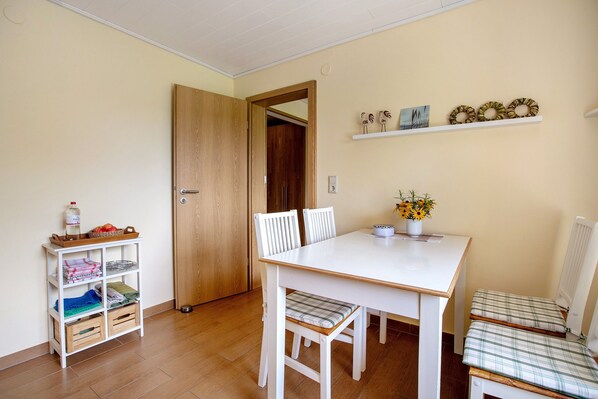 A dining area with a white table, two chairs, a small shelf with items, and a wooden door. - Apartment 'Ferienwohnung Am Eiderdeich' with Private Terrace, Shared Garden and Wi-Fi (Delve)