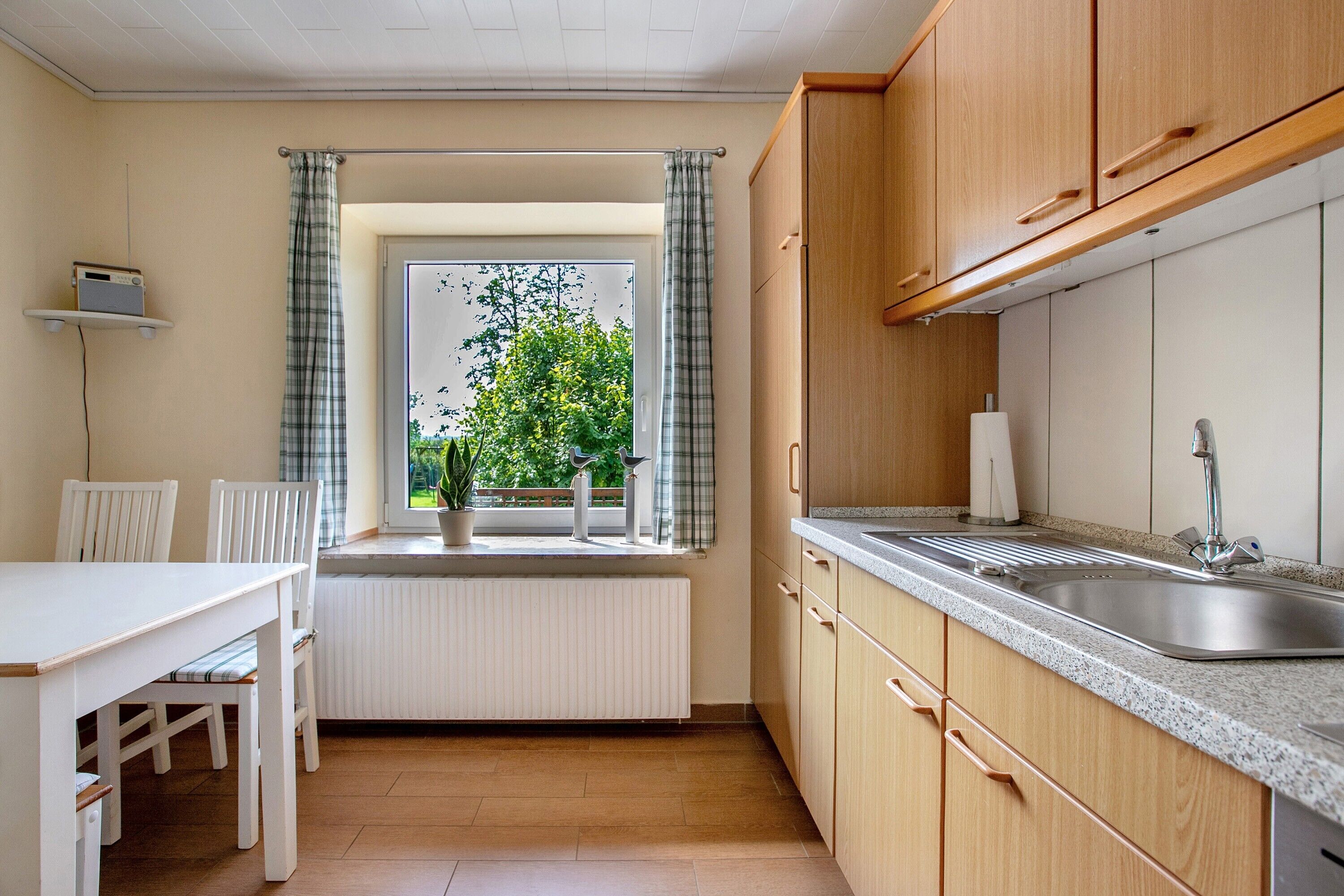 Une cuisine de taille réduite, avec une fenêtre, un radiateur blanc, des placards en bois et un évier.