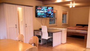 A room with a wooden dining table, two chairs, a desk with a chair, a bed, a TV mounted on the wall, and a patterned rug.