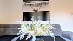 Ett vardagsrum med en grå soffa, en tavla med ett rådjur som hänger på väggen och en vas med vita blommor.