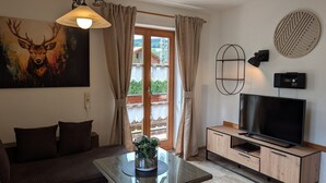 A living room with a sofa, a TV on a wooden stand, a painting of a deer, and a window with curtains. - Apartment 'Wohnung 1' with Mountain View, Balcony and Wi-Fi (Mittenwald)