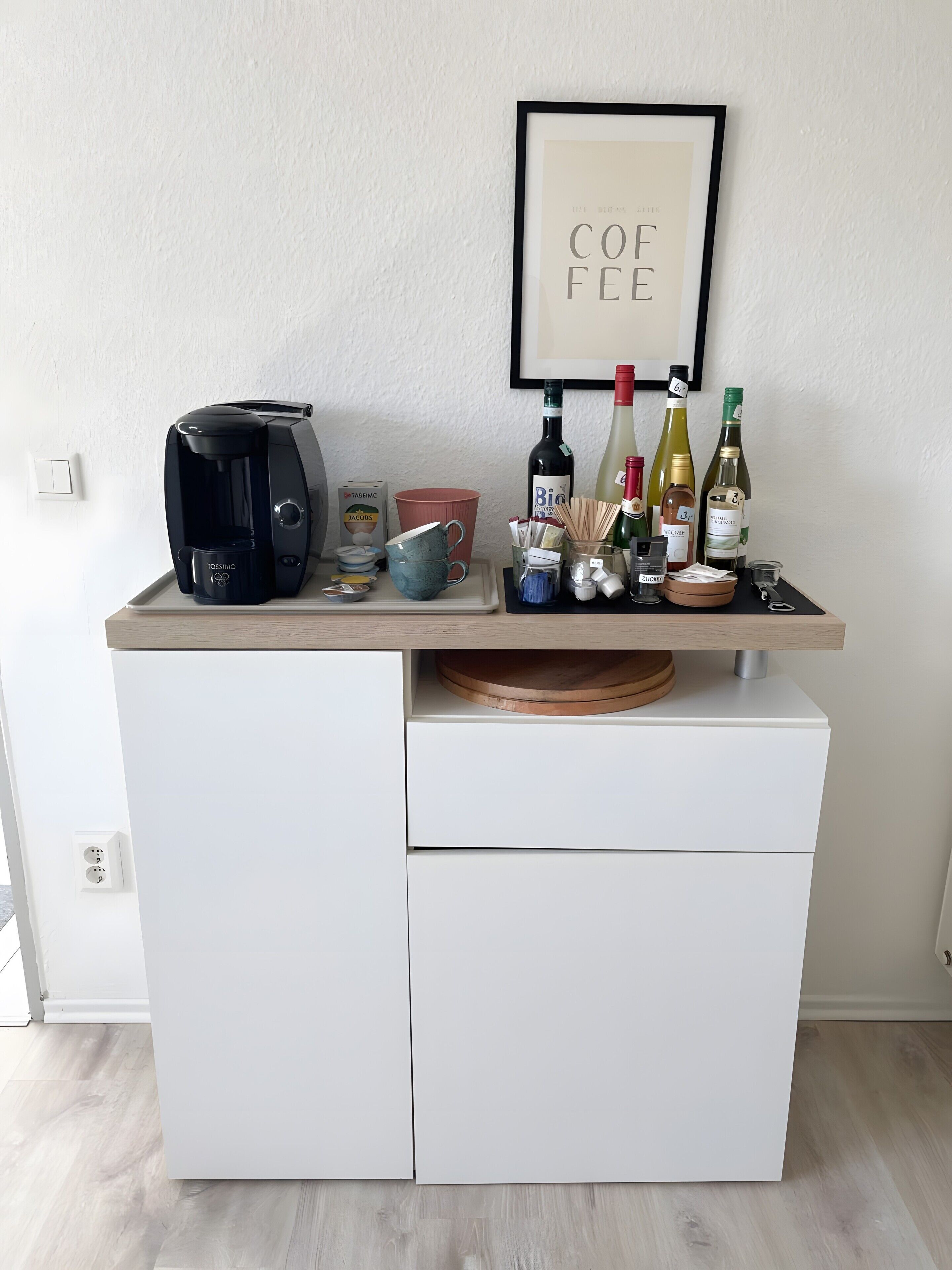 A kitchenette with a coffee maker, mugs, wine bottles, and a framed sign on the wall.