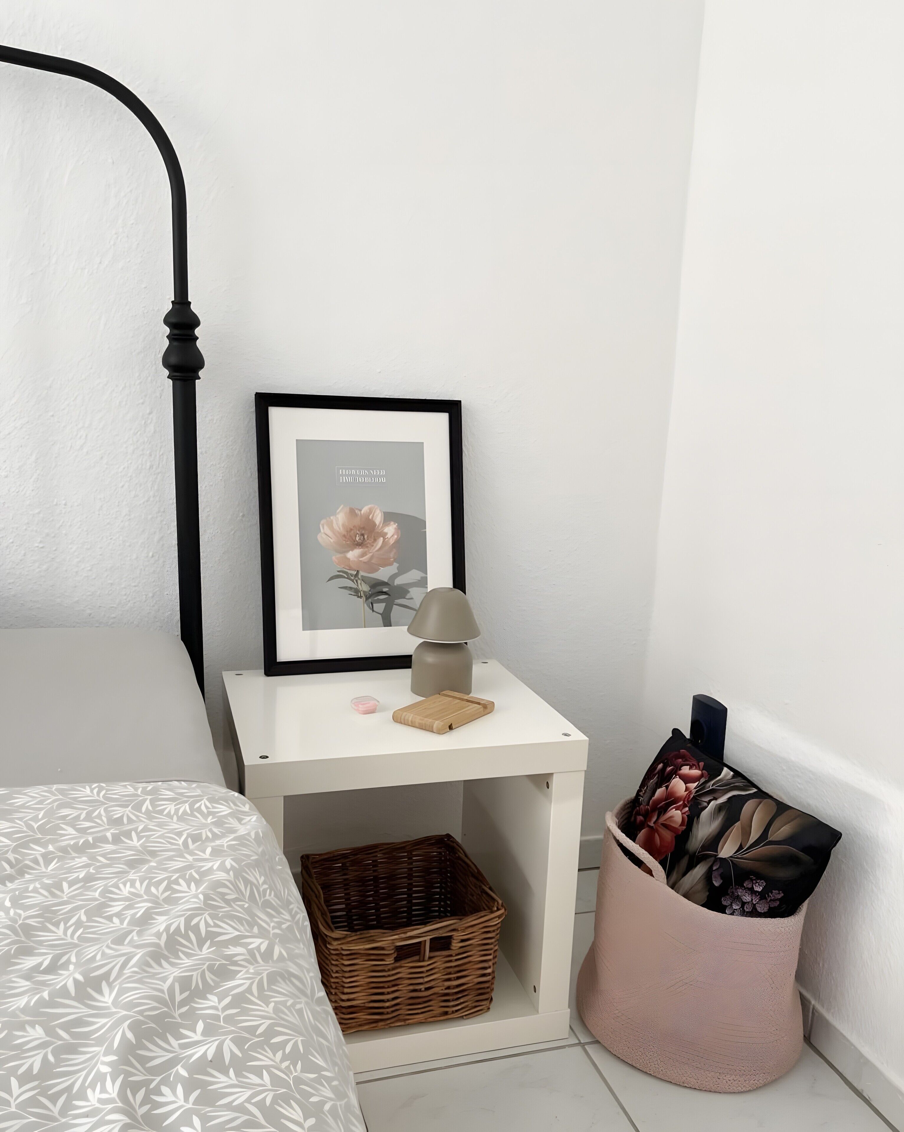 Un dormitorio ordenado con cama, mesita de noche con lámpara, un cuadro enmarcado, una cesta pequeña y un recipiente de almacenamiento.