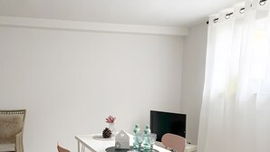 Ein schlicht eingerichteter Raum mit einem weißen Tisch, zwei Stühlen, einem kleinen Teppich, einem Fernseher und einem Fenster mit weißen Vorhängen.