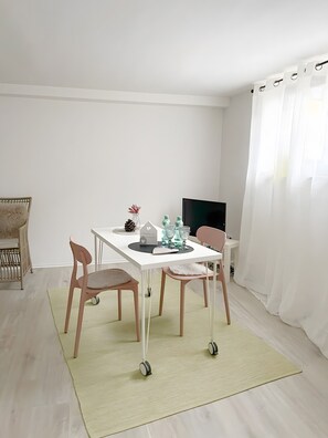 A minimalist room with a white table, two chairs, a small rug, a television, and a window with white curtains.
