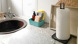 Ein Badezimmer mit weißem Waschbecken, einem Spiegel, einem gerahmten Schild über dem Spiegel, einem Regal mit Tassen und Wasserkocher sowie einem Toilettenpapierhalter.