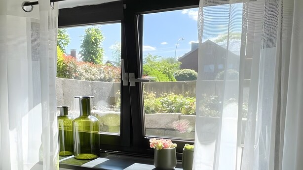 Ein Zimmer mit Fenster, weißen Vorhängen und Blick auf Grünfläche.
