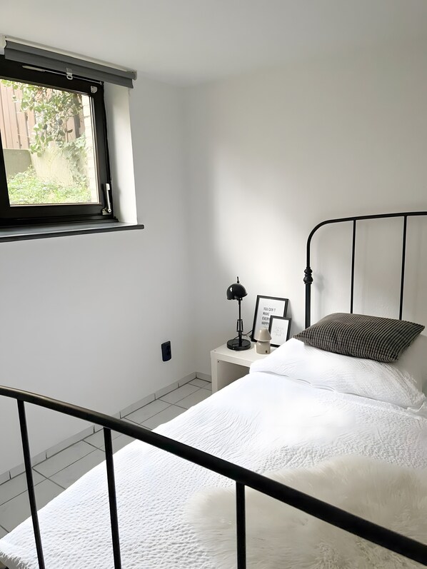 Ein Schlafzimmer mit einem Bett, einem Nachttisch, einer Lampe, einem Kissen, einem gerahmten Bild und einem Fenster mit Blick auf Grünflächen.