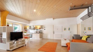 A spacious living area with a flat-screen TV, a sofa, a dining table, and a kitchen in the background.