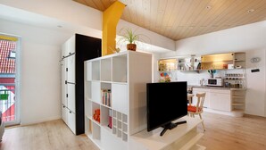 A modern living room with a flat-screen TV, white shelving, a potted plant, and a kitchen area with white cabinets and a microwave.