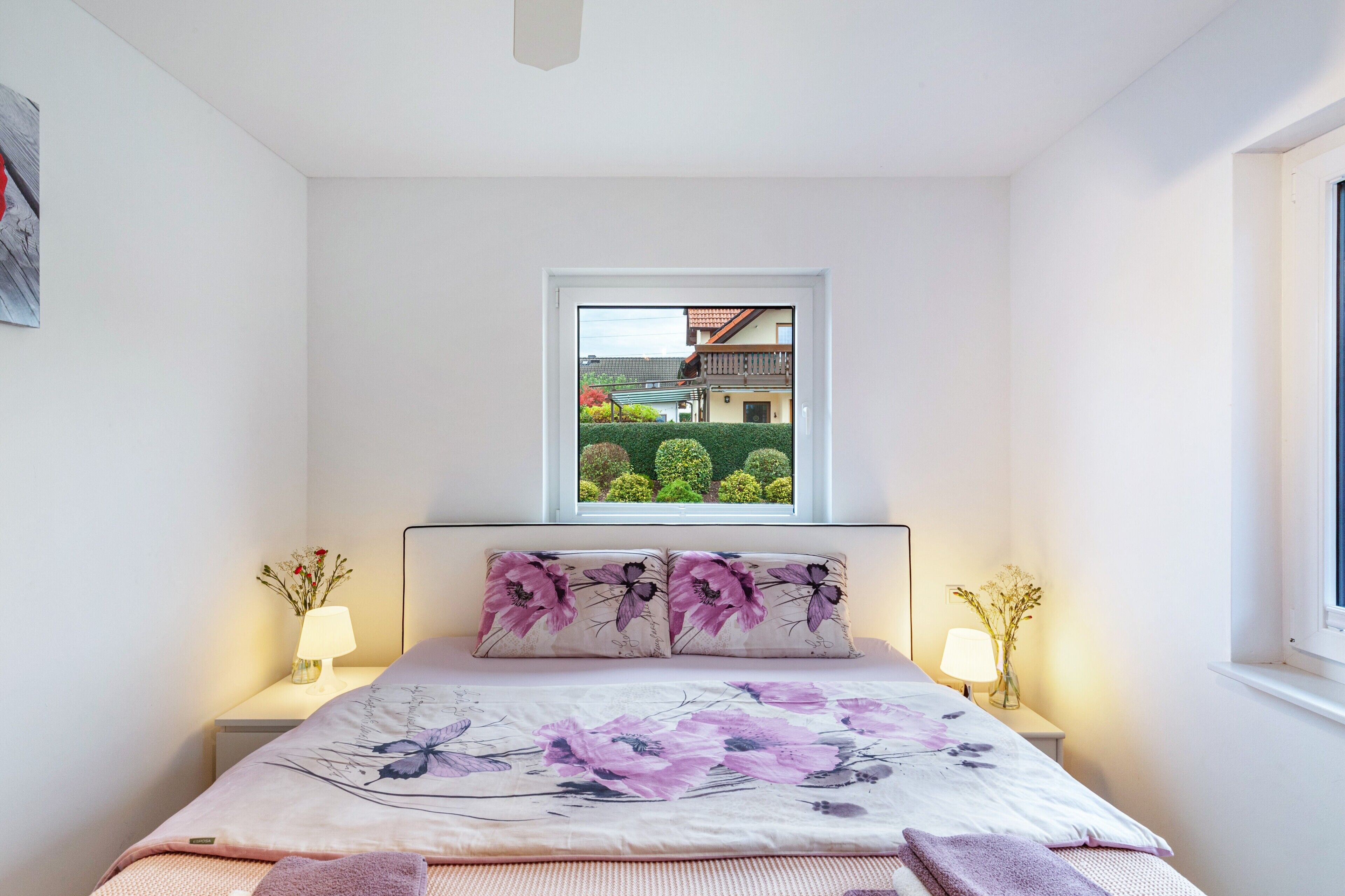 Un dormitorio con cama, dos lámparas de noche, una ventana con vistas a un edificio y follaje, y un cojín decorativo.