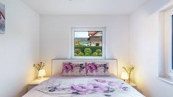 Un dormitorio con cama, dos lámparas de noche, una ventana con vistas a un edificio y follaje, y un cojín decorativo.